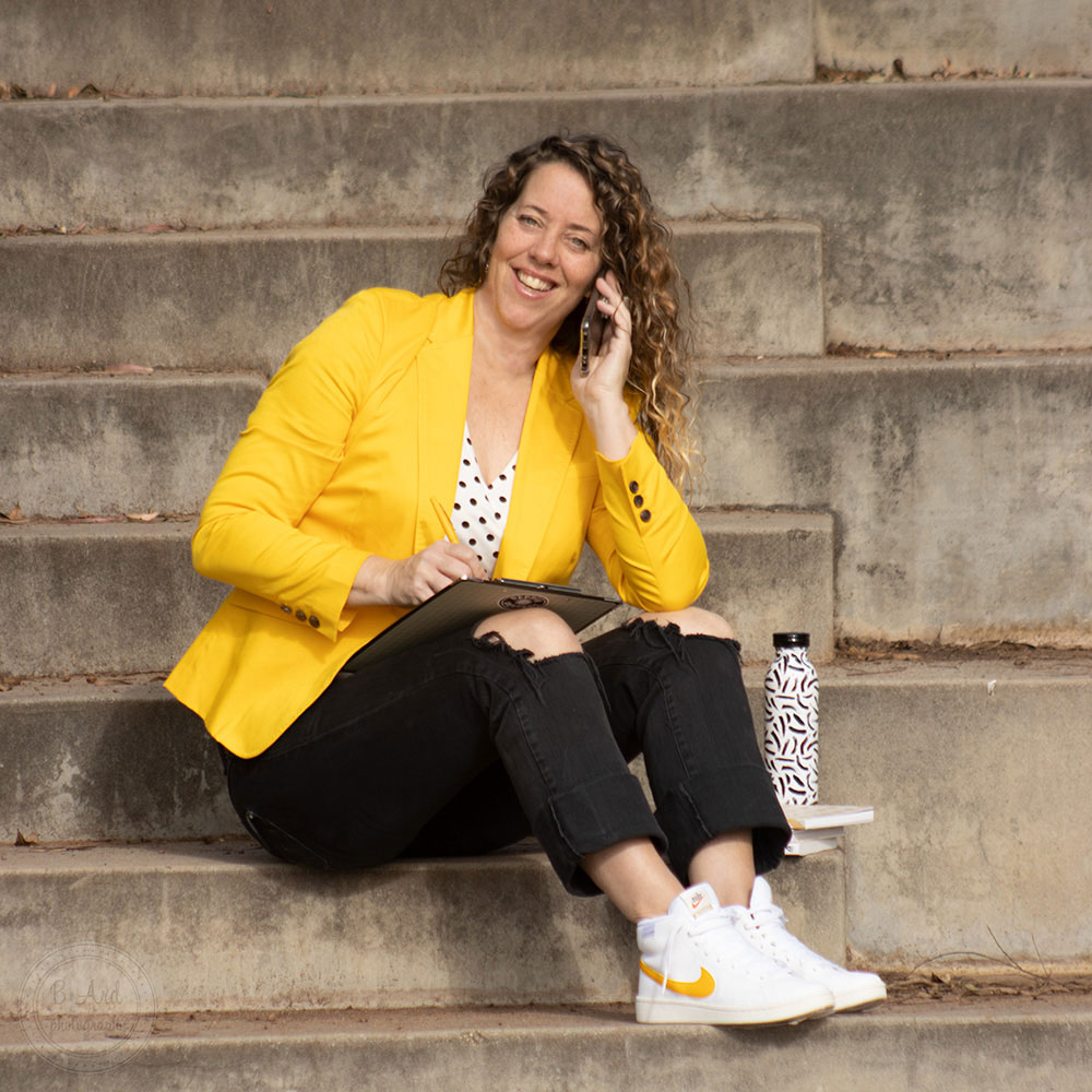 A lady, with long curly hair is sitting on a set of stairs while talking on her phone and making notes about remarks.