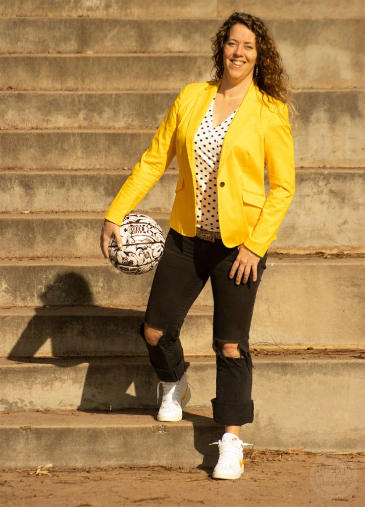 A young lady at the lowest step of a set of stairs, wears a bright yellow business jacket, black jeans and basketball shoes. She is holding a basketball on her right hip.