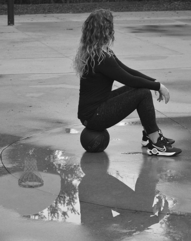 A female basketball coach is sitting on a basketball in the middle of an empty outdoor basketball court.