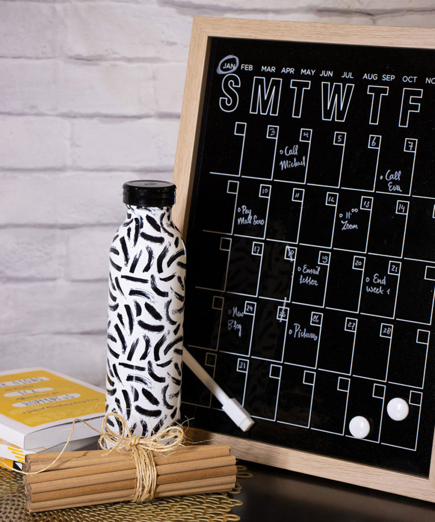 A wood framed erasable calendar reflects a filled-in schedule. As decorative items, a white bottle with black stripes, a set of books and wooden pencils are laying in front of the framed calendar.