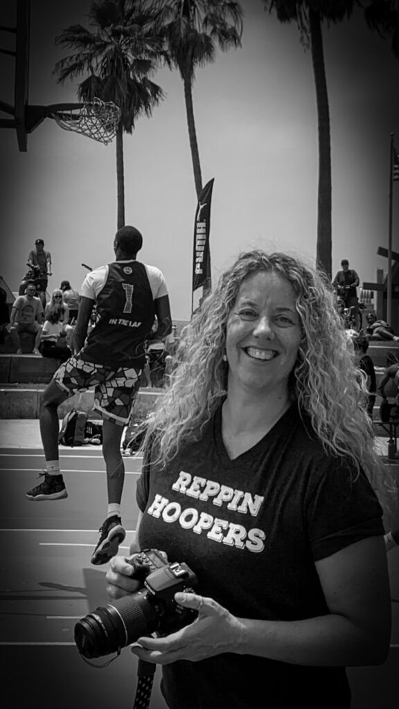 A female sports photographer is seenon the sidelines of an outdoors basketball game holding a camera in her hands.